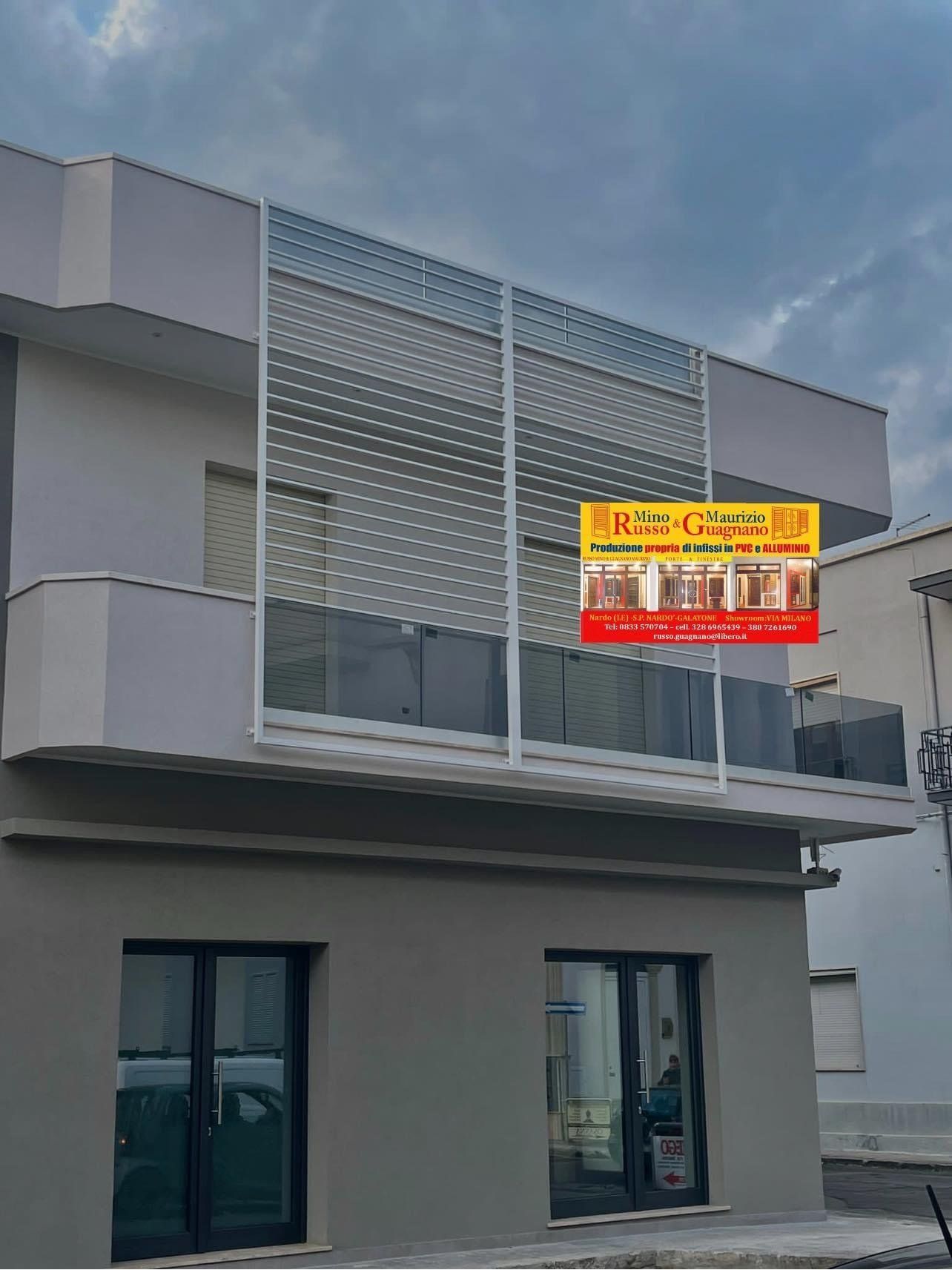 Edificio a due piani con facciata grigia, balcone in vetro, schermo in metallo bianco e insegna aziendale.