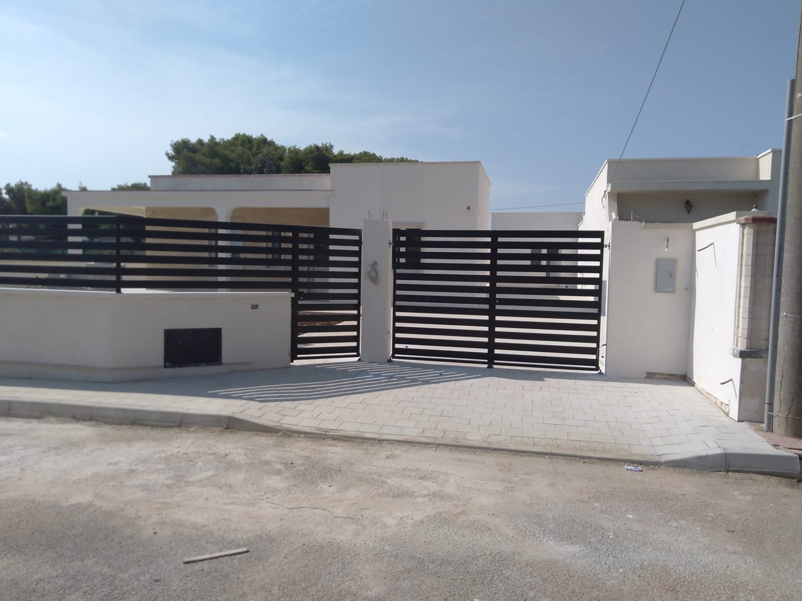 Casa bianca con cancello orizzontale nero, vialetto in cemento e cielo azzurro.