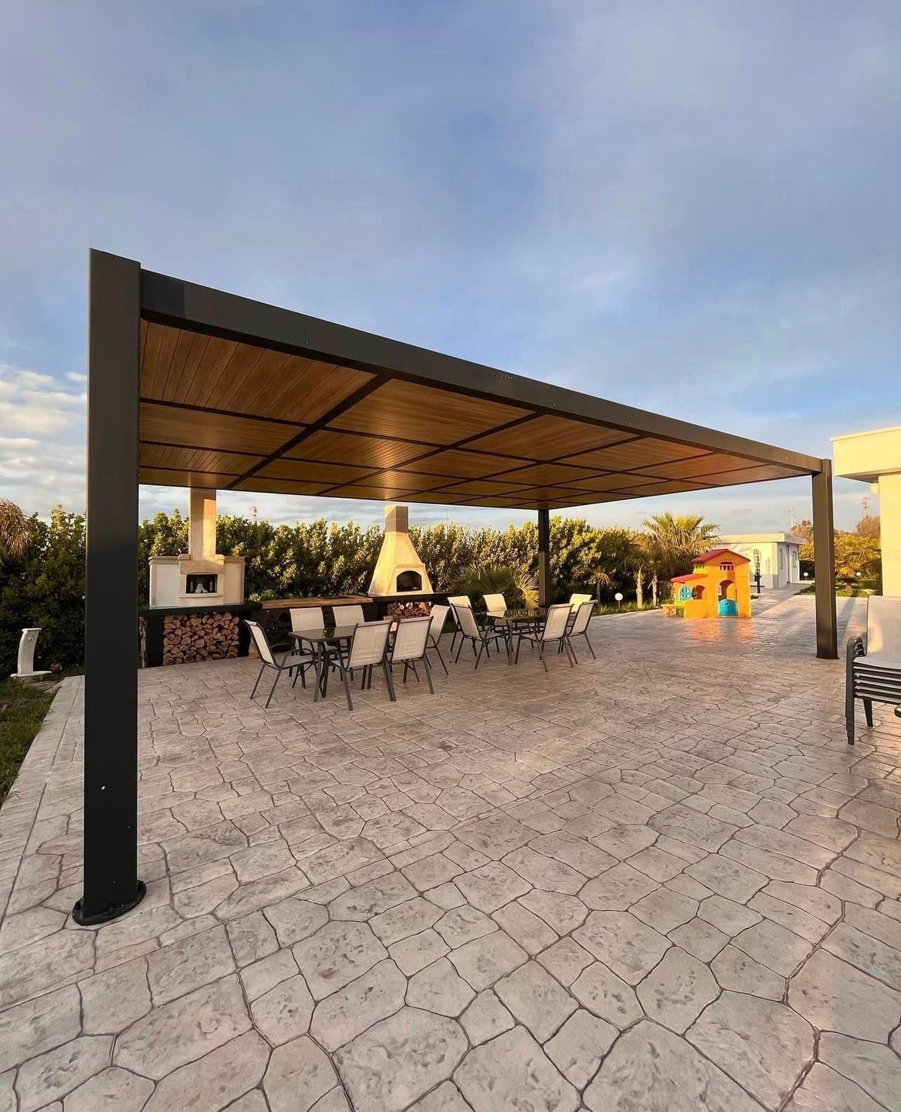 Zona pranzo all'aperto con pergola rivestita in legno, forno per pizza, tavoli e sedie su un patio in pietra.