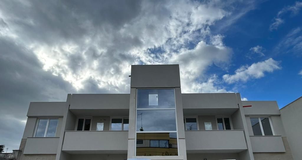 Moderno edificio grigio chiaro sotto un cielo parzialmente nuvoloso, con vetrate riflettenti al centro.