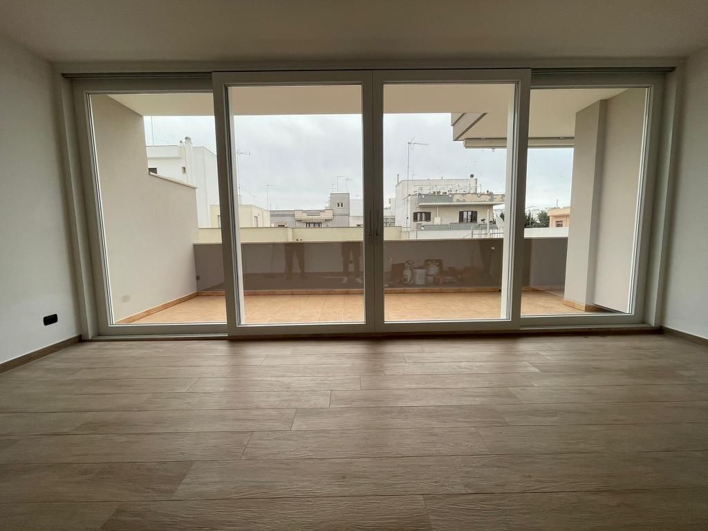 Stanza vuota con pavimento in legno, grandi porte a vetri che si aprono su una terrazza con vista sulla città.