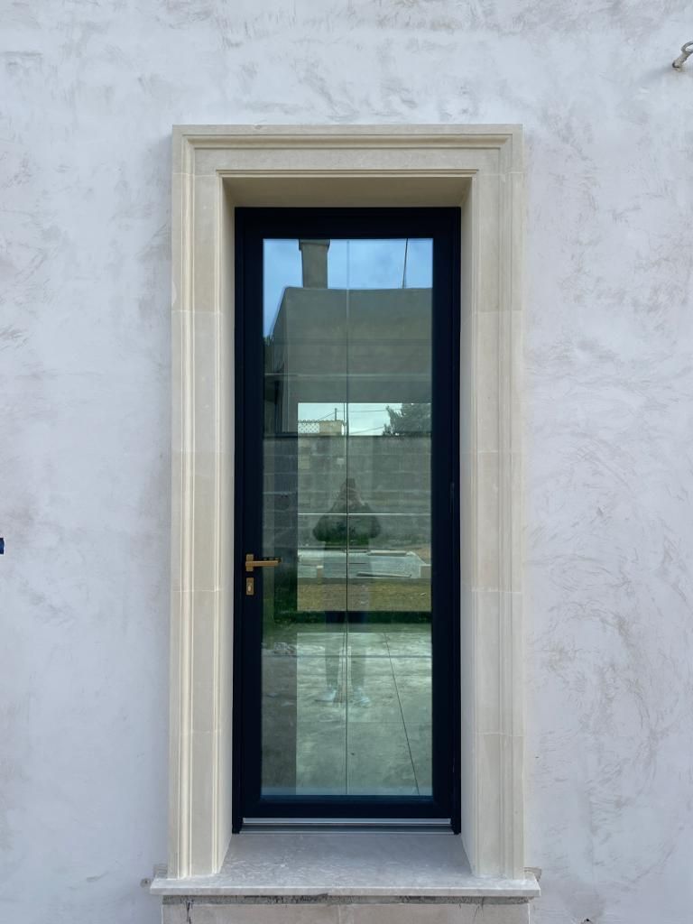 Una porta in vetro nero con cornice e finestra in pietra calcarea, inserita in una parete in stucco bianco.