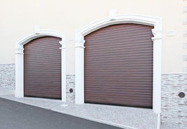 Due porte da garage avvolgibili marroni con telai ad arco bianchi, nella facciata di un edificio con dettagli in pietra.