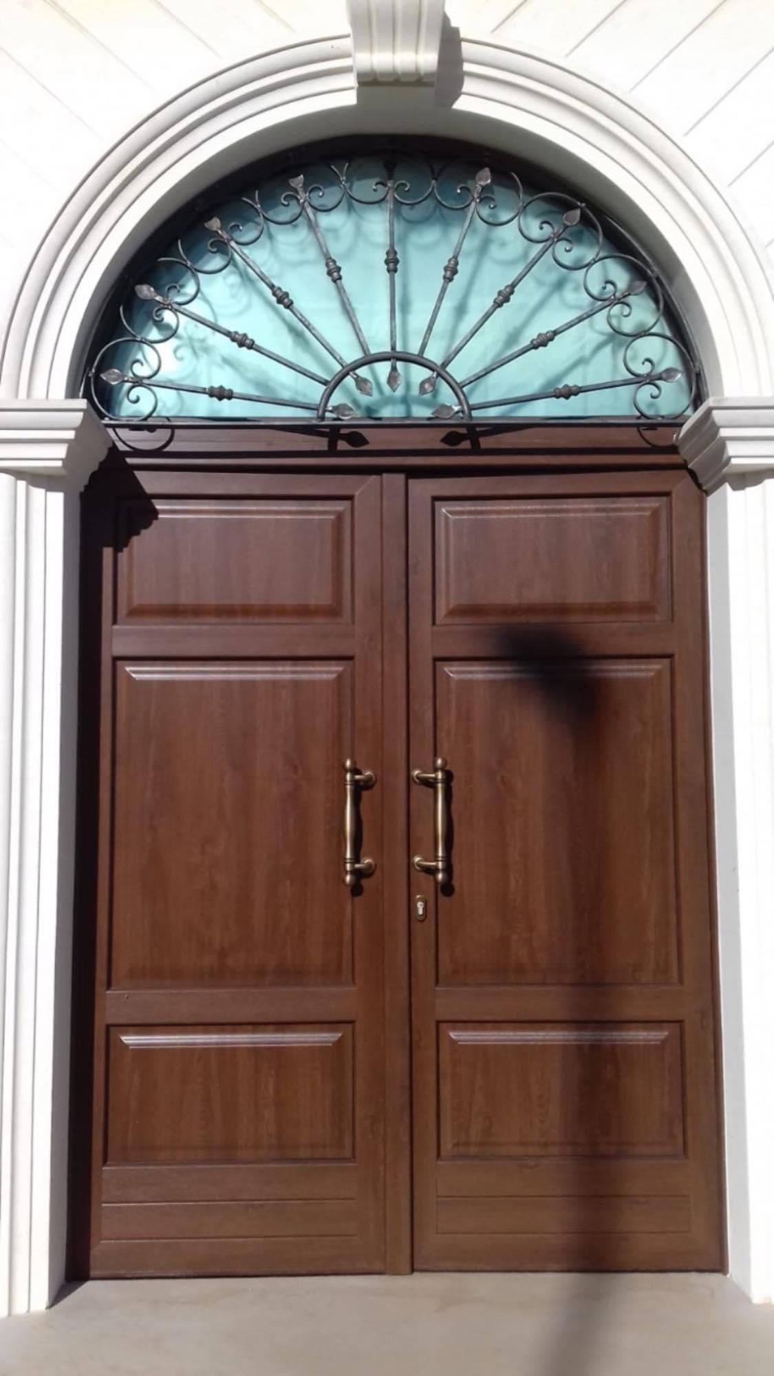 Porte doppie marroni con parte superiore arcuata e decorazioni in ferro battuto, inserite nella facciata in pietra bianca.