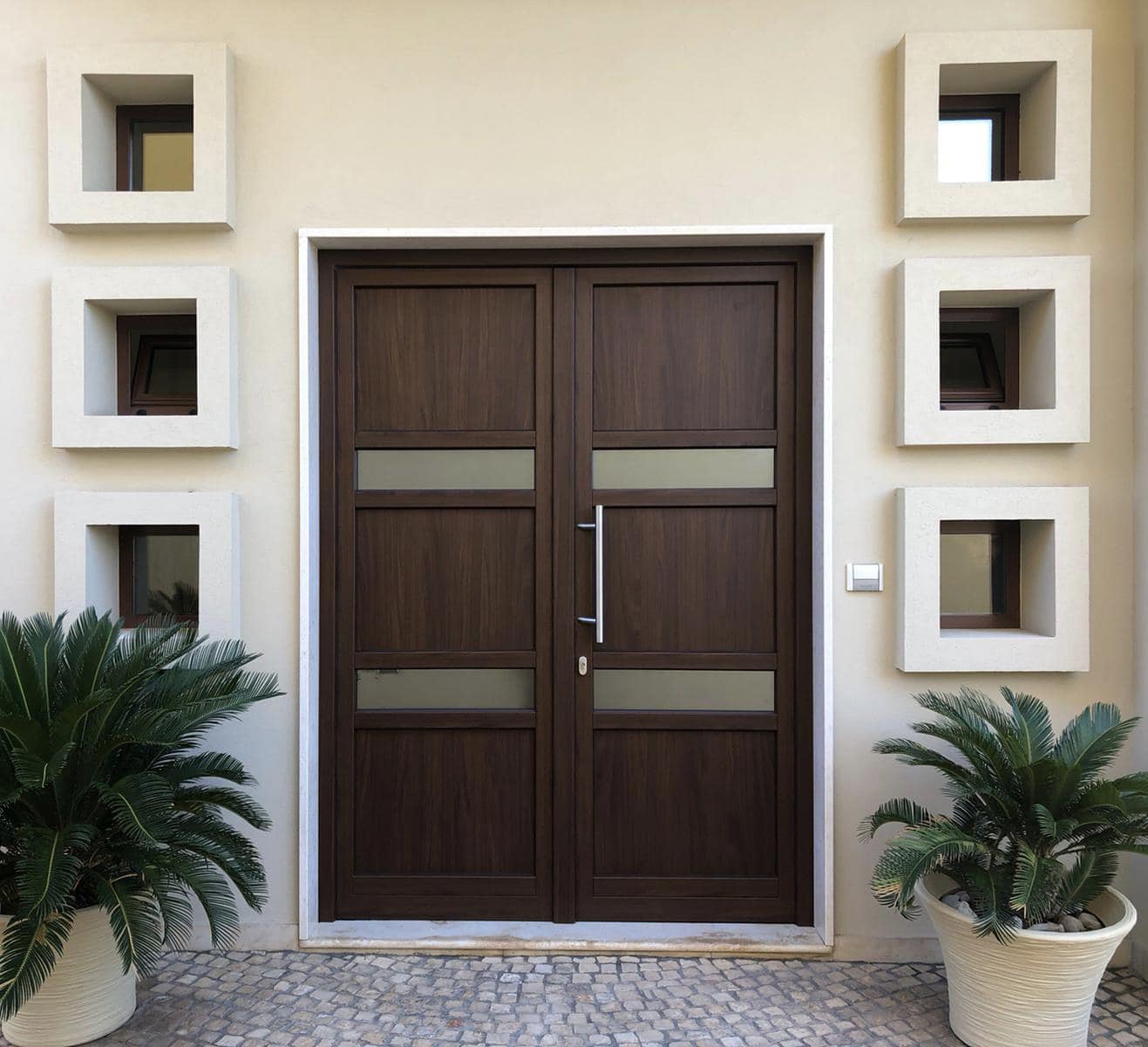 Doppie porte marroni con pannelli in vetro, fiancheggiate da finestre quadrate e piante in vaso.