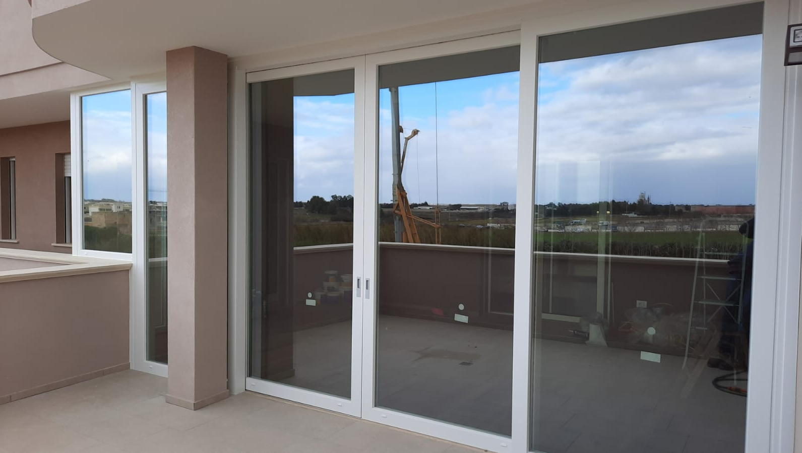 Porte scorrevoli in vetro con cornici bianche su un balcone, che riflettono il cielo e il paesaggio.