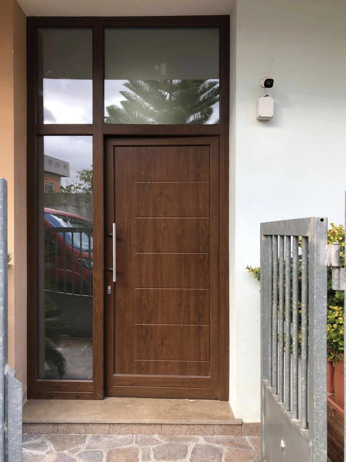 Porta d'ingresso marrone con finestra laterale e sopraluce; ambiente esterno.
