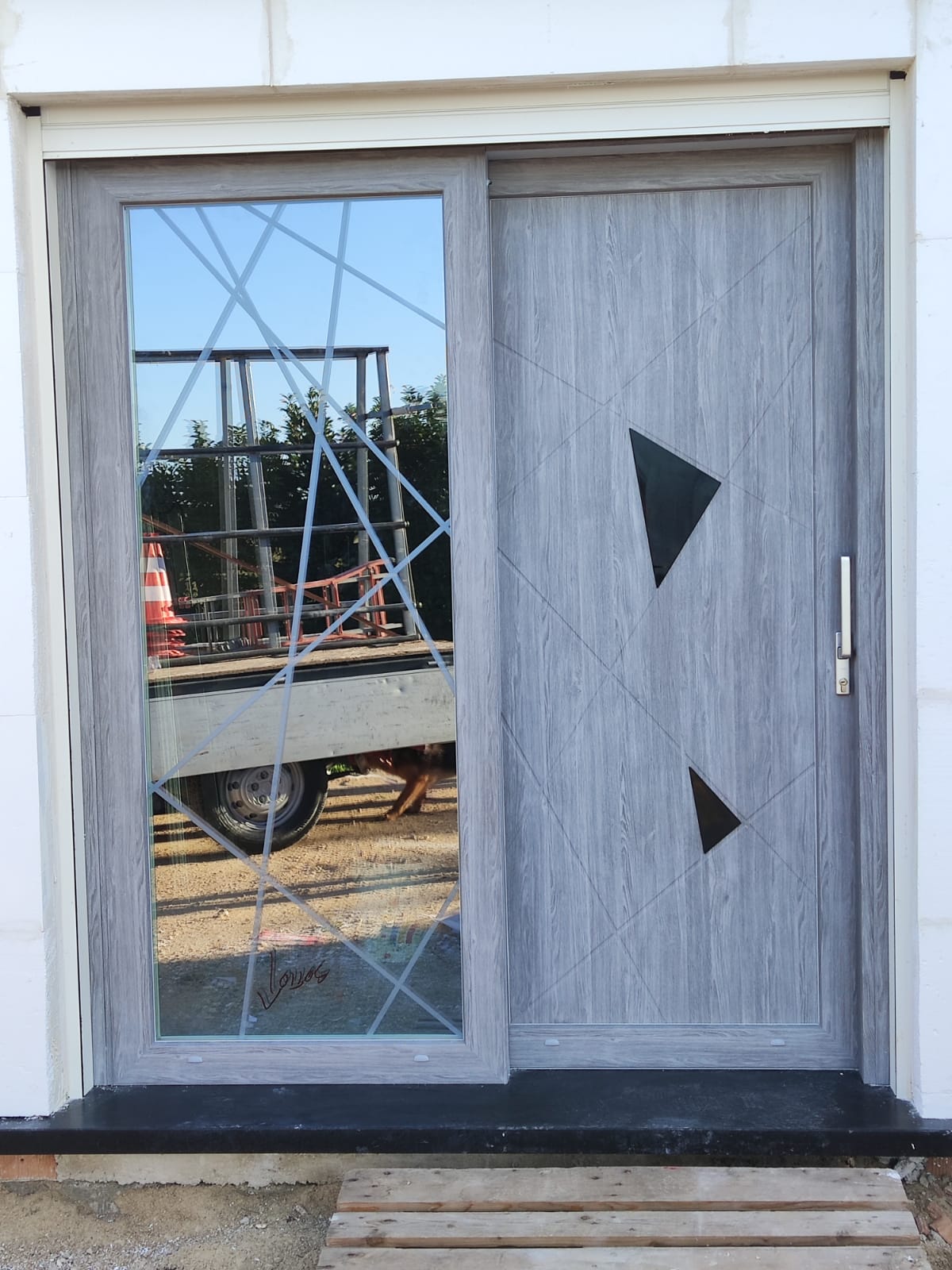 Porta scorrevole grigia con finestra geometrica che riflette un cantiere edile; 