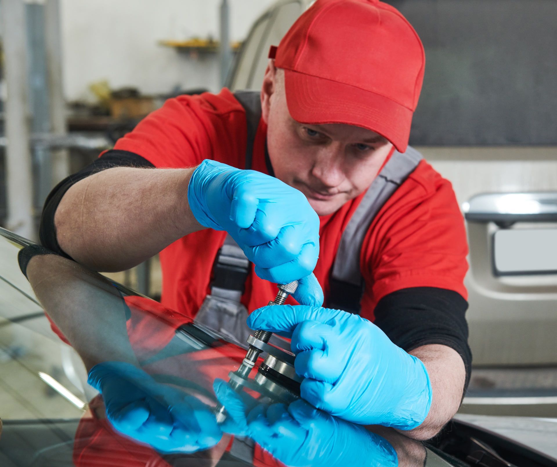 Mechanic in red shirt and cap repairing a windshield, wearing blue gloves.