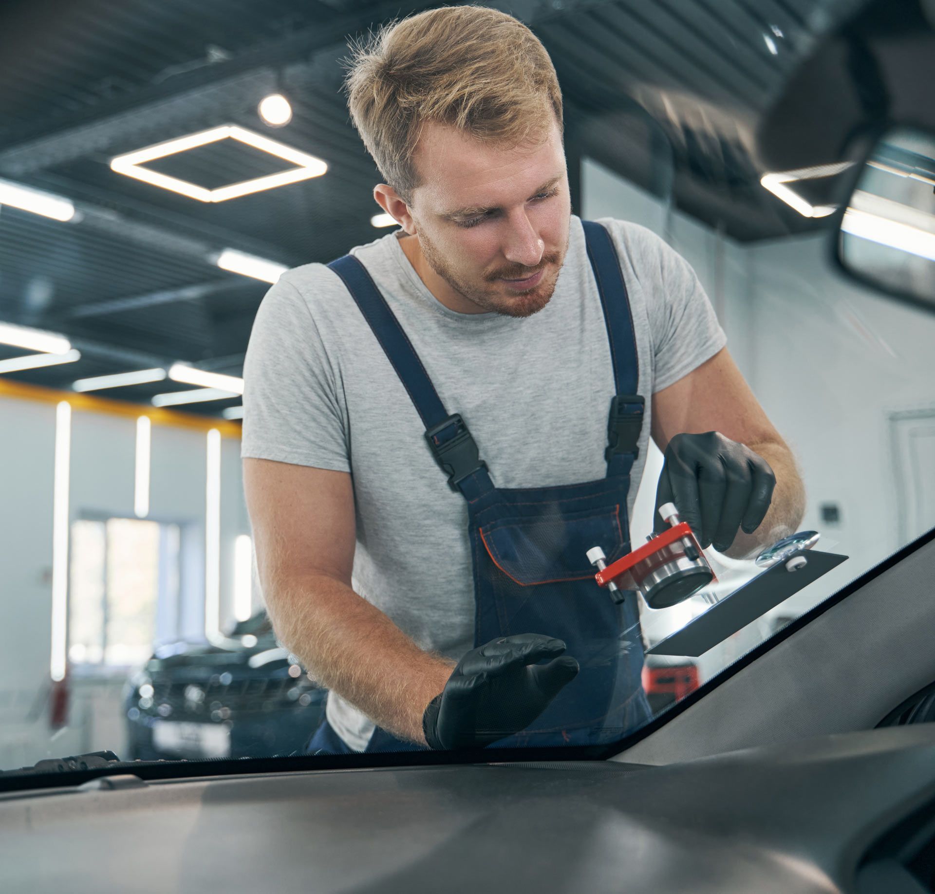 Mechanic in overalls repairing a car windshield. He is wearing gloves and holding a tool in a garage.