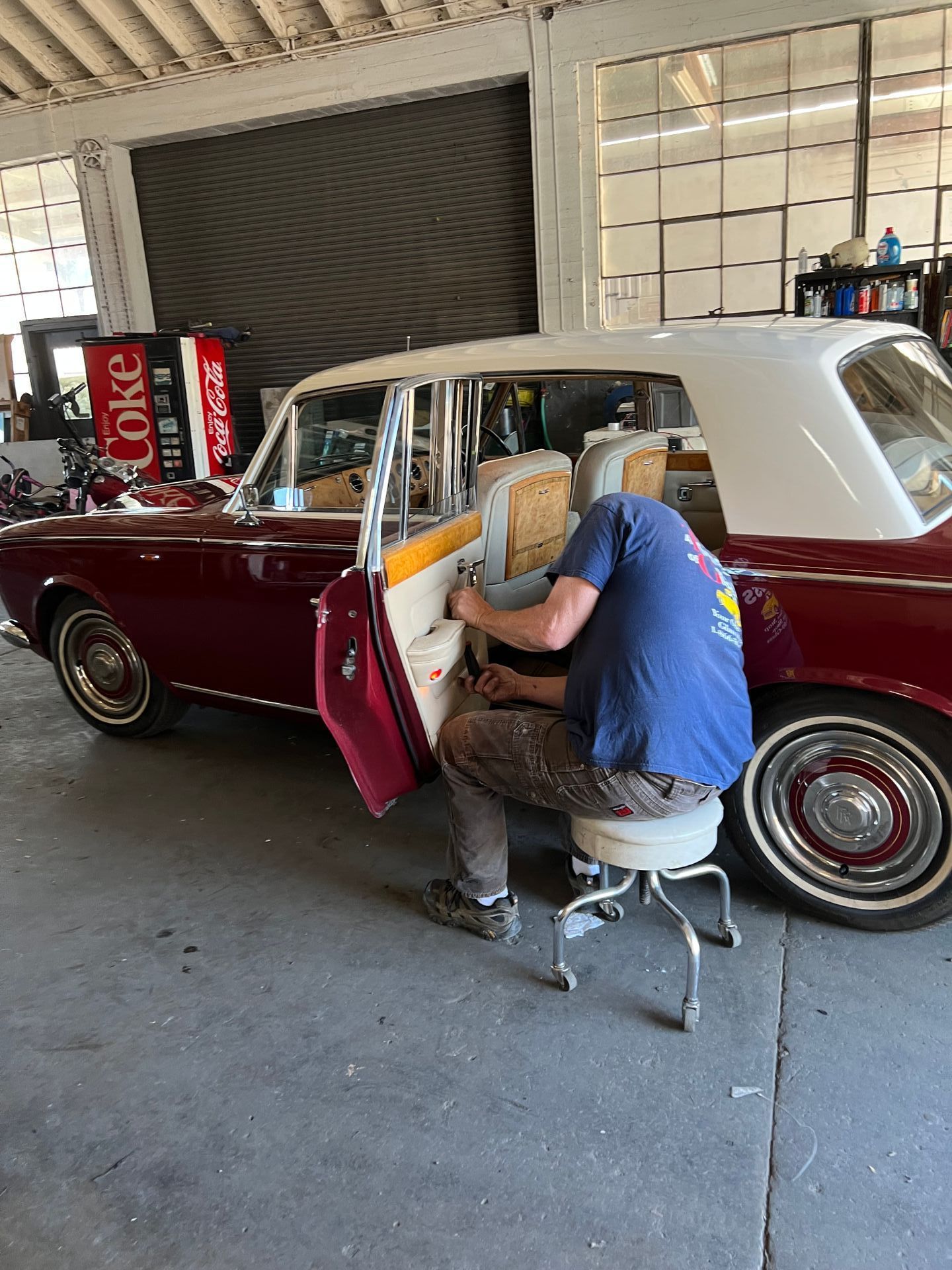 A person works on the interior of a maroon and white vintage car in a garage.