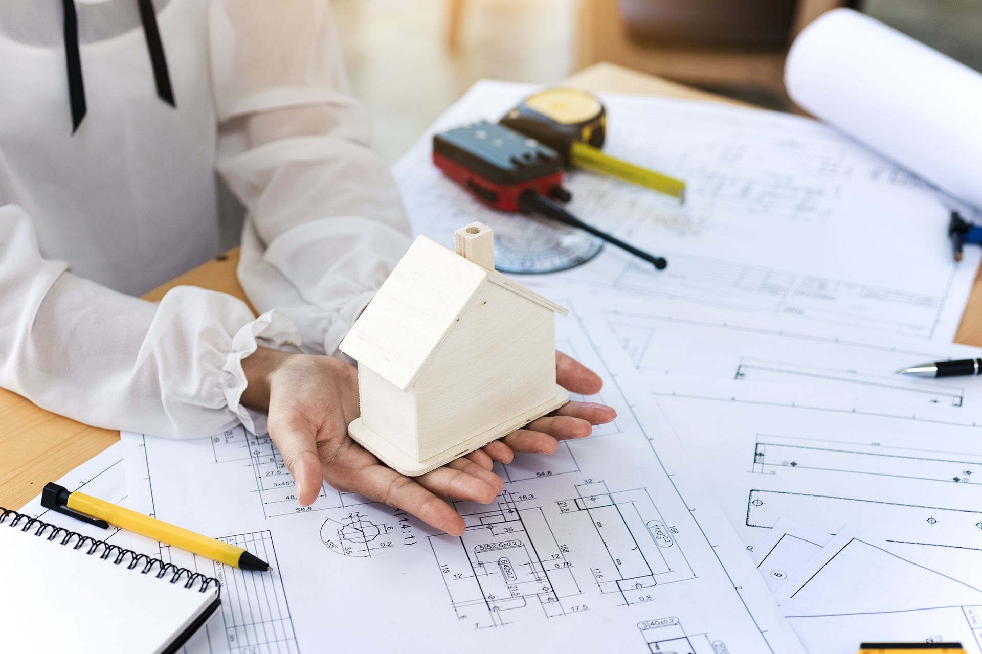 Person holding a small house model above architectural blueprints on a desk.