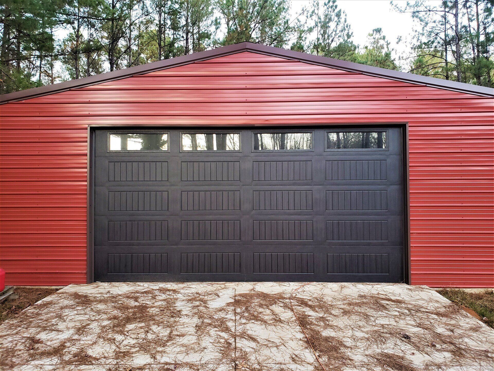 A red garage with a black garage door is sitting in the middle of a forest.
