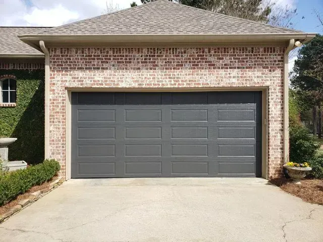 A dark gray, four-panel residential garage door set into a brick exterior wall above a concrete driveway.