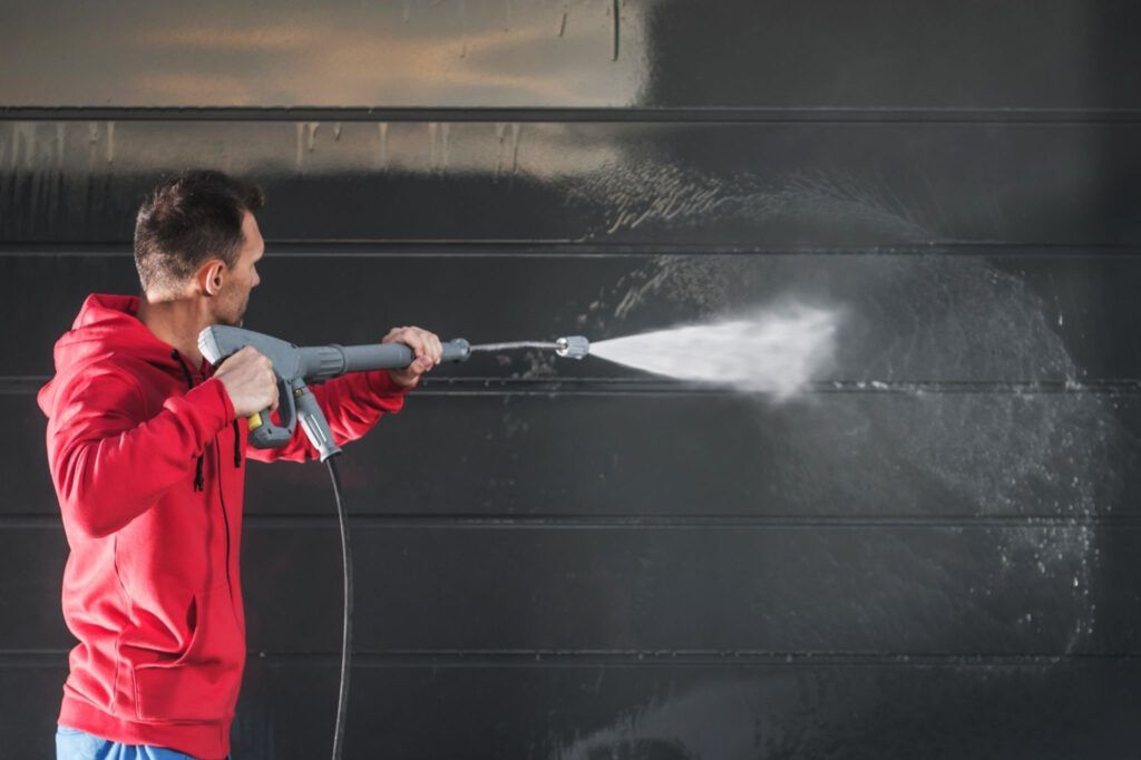 A man is using a high pressure washer to clean a garage door.