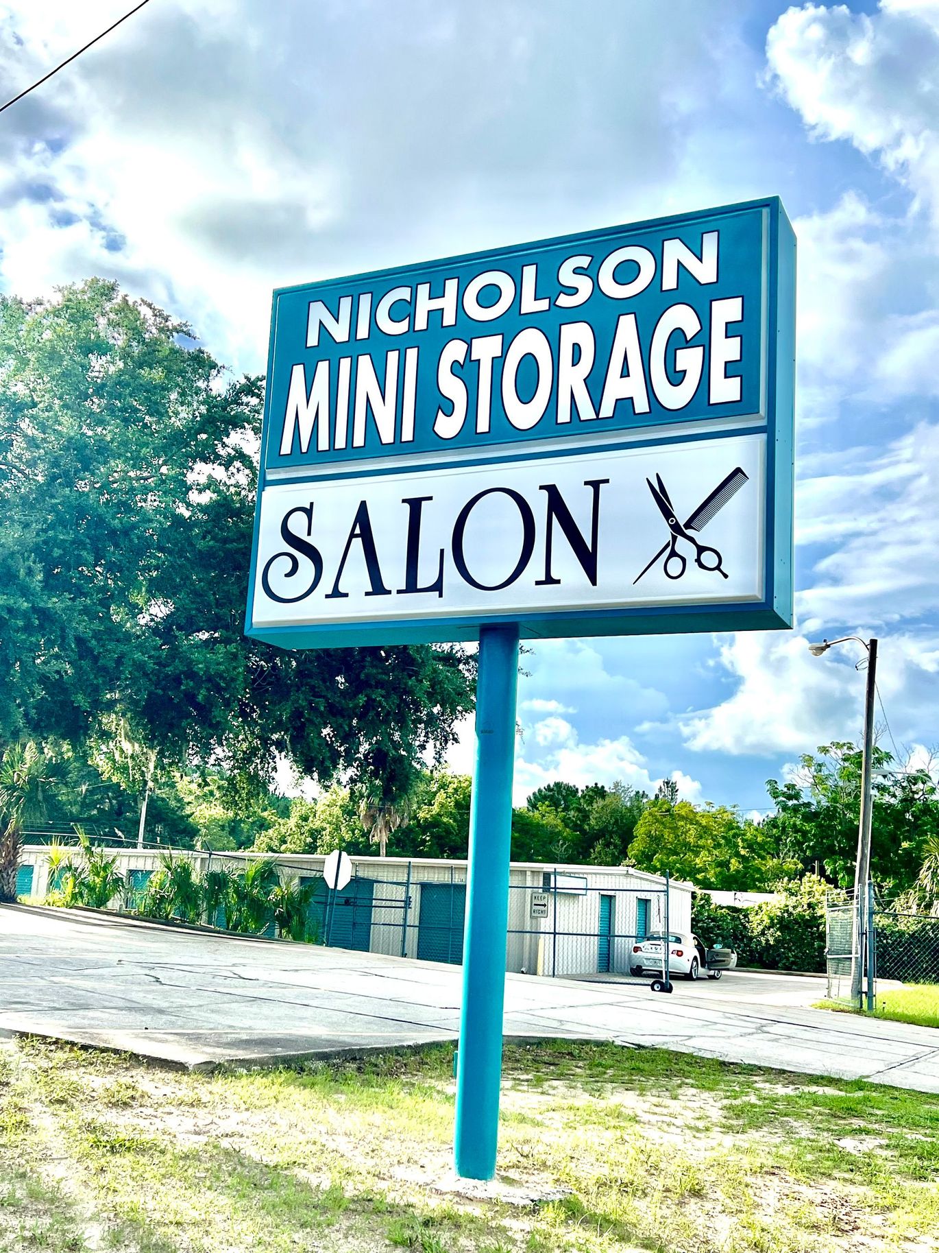 Sign for Nicholson Mini Storage and Salon, blue and white, near storage units under a cloudy sky.
