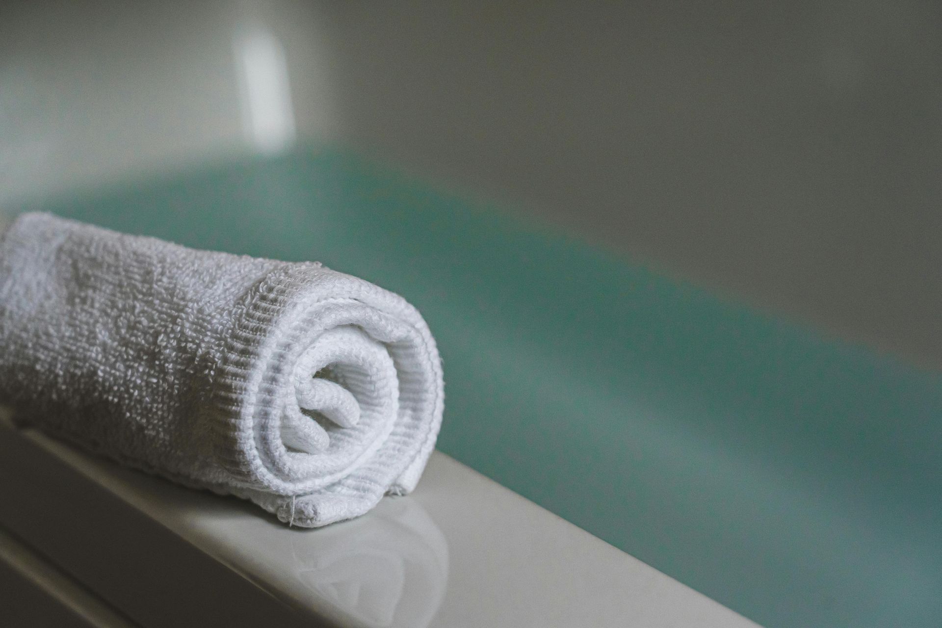 Rolled white towel on the edge of a bathtub filled with light blue water.