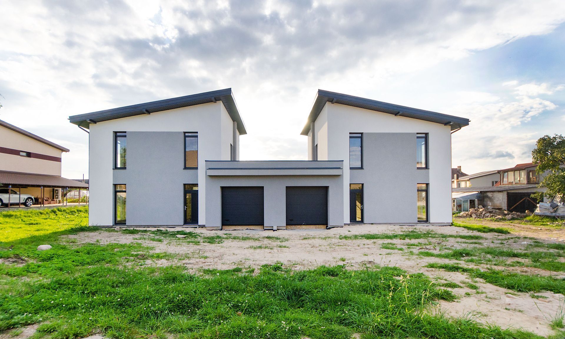 Modern white duplex house with dark roof and garage, viewed from a grassy front lot.