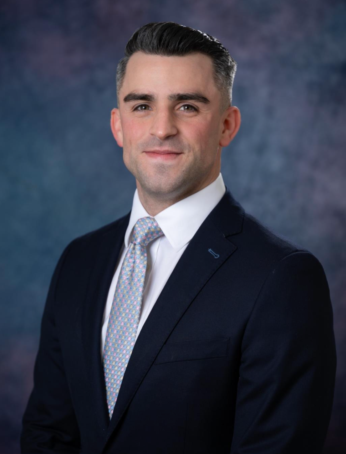 A smiling man in a suit and tie poses for a professional portrait against a blurred background.