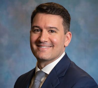 Smiling man in a navy suit and patterned tie, against a blurred blue background.