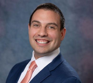 A smiling man with fair skin wearing a blue suit and coral tie, set against a blurry blue and purple background.