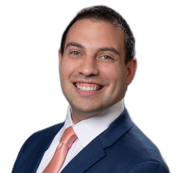 A smiling man with fair skin wearing a blue suit and coral tie, set against a blurry blue and purple background.