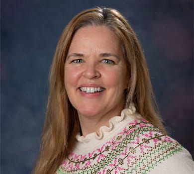 A woman with long blonde hair smiles, wearing a patterned sweater, against a blue background.
