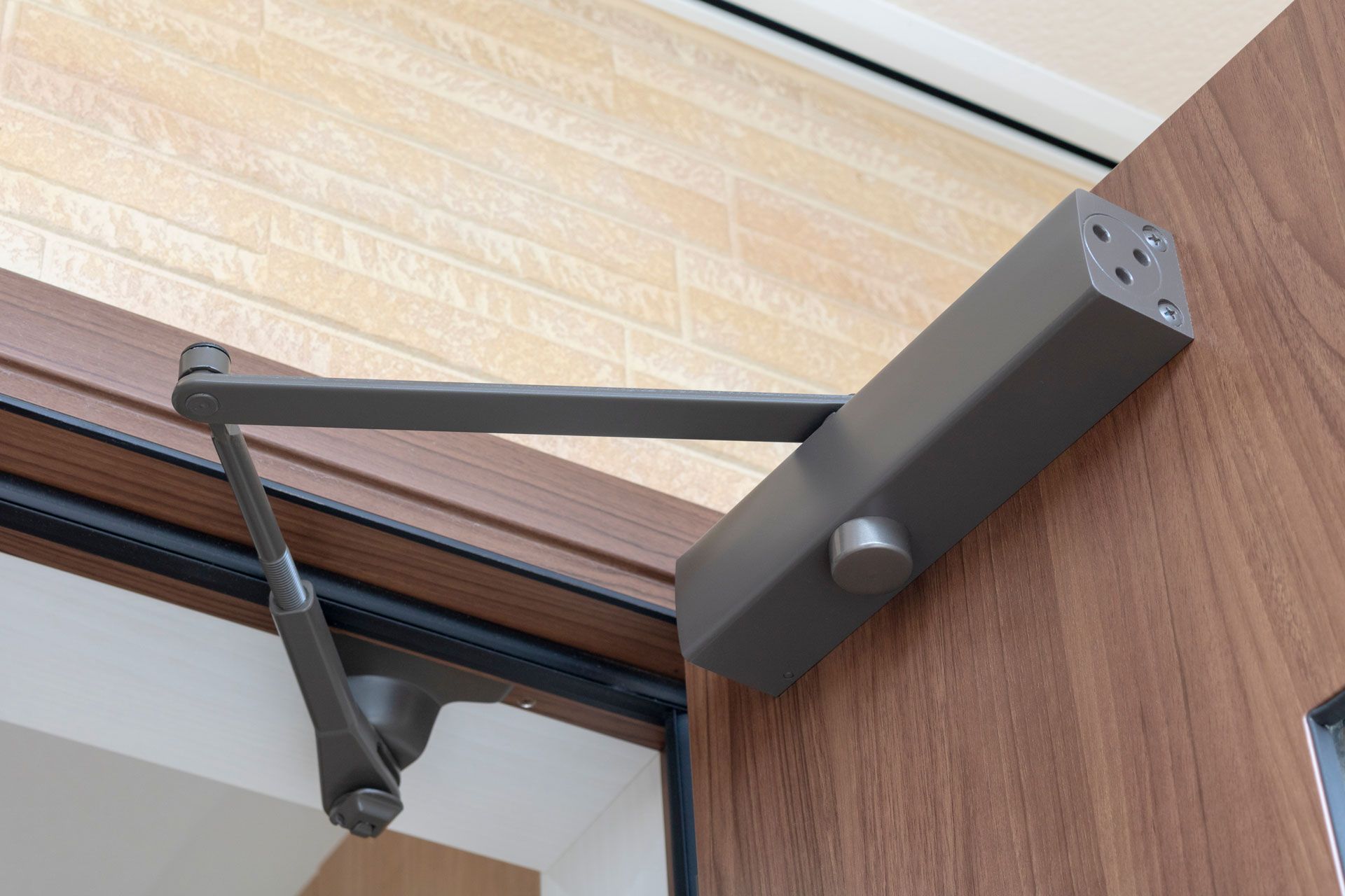 Door closer mechanism mounted on a brown wooden door, with a gray arm and a light beige wall in the background.