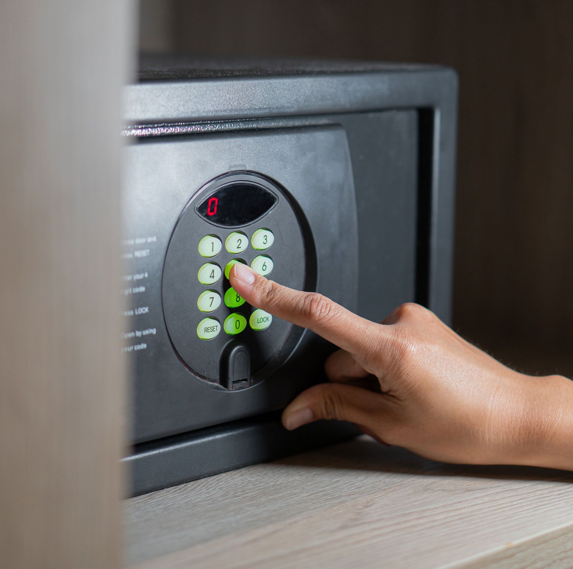 Person's hand pressing a button on a black digital safe, set on a wooden shelf.