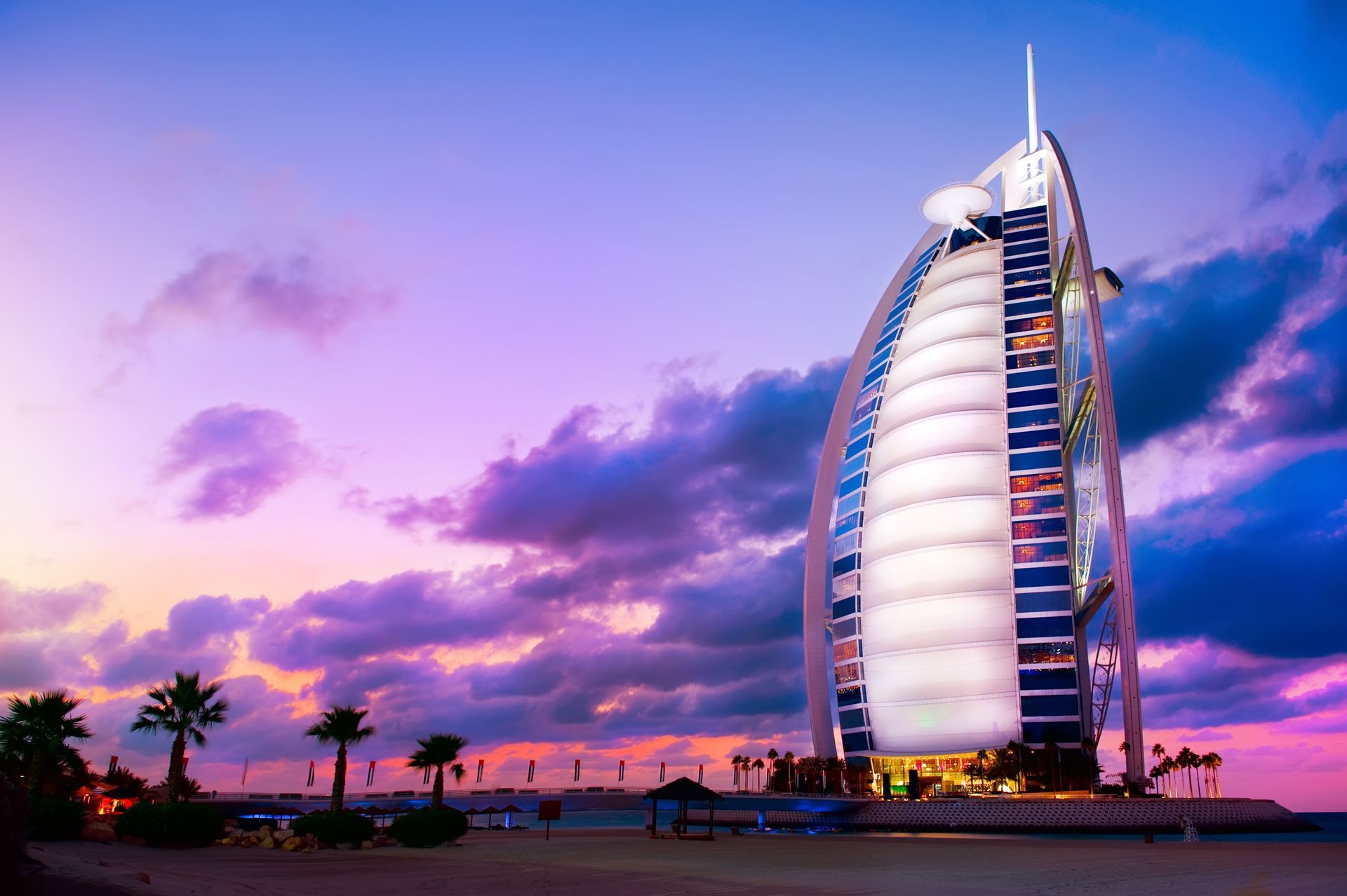 Tall sail-shaped hotel by the waterfront at sunset, with palm trees and pink-purple clouds.
