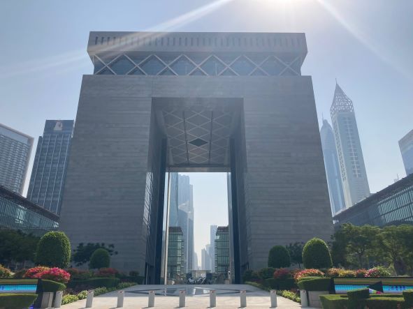 Modern gray gateway building in bright sunlight, framed by city skyscrapers and landscaped street-level greenery