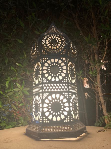 Glowing ornate metal lantern on a table outdoors at night, surrounded by plants