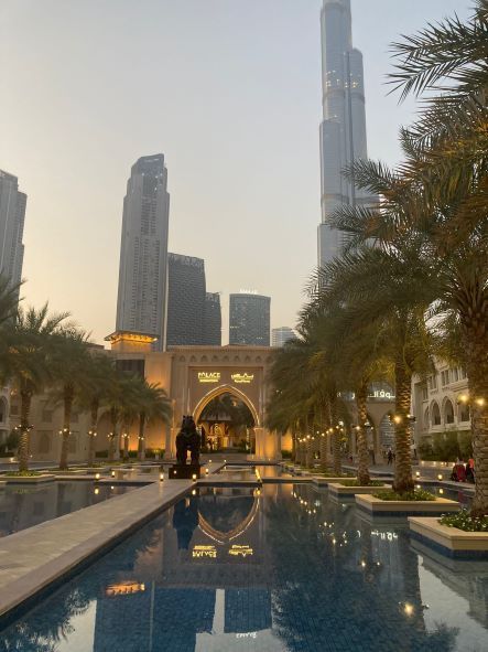 Dubai skyline reflected in a pool at sunset, with palm trees and a lit archway in the foreground