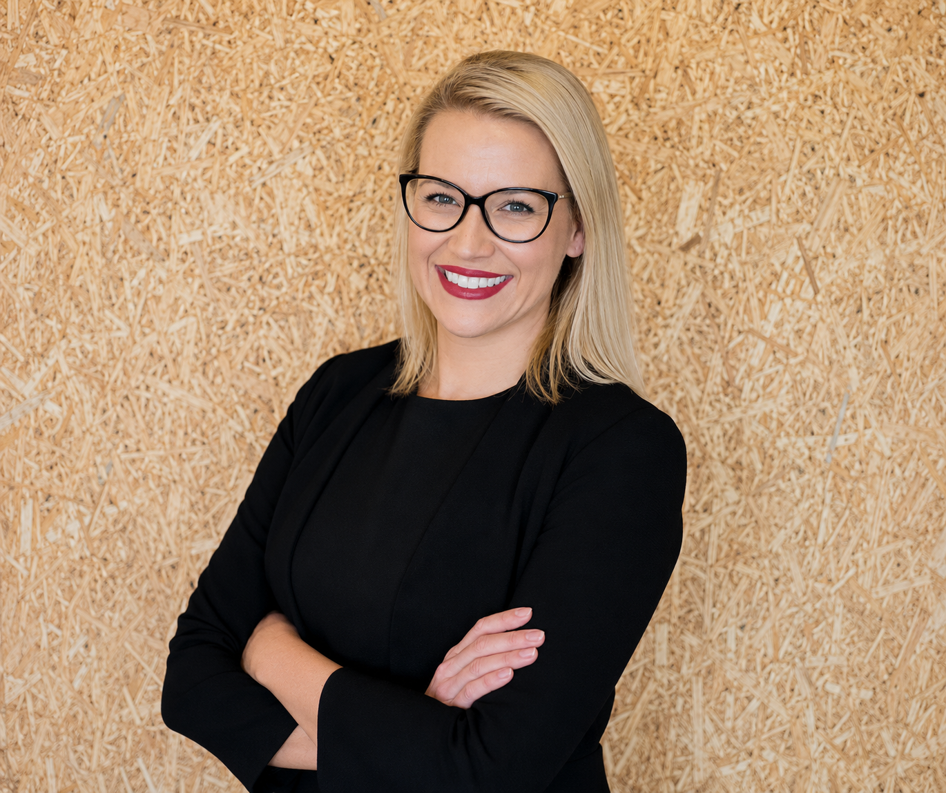 Smiling person with glasses and blonde hair, wearing a black top, standing with arms crossed against a wood wall