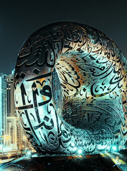 Large reflective Arabic calligraphy sculpture lit at night in a city plaza, with buildings behind it