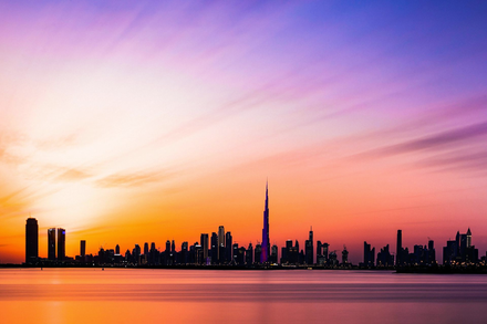 Silhouette of a city skyline at sunset with a tall central tower and pink-orange sky over calm water