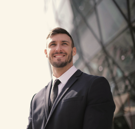 Smiling man in a dark suit standing outdoors near a glass building