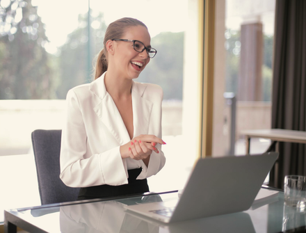 Smiling woman in a white blazer sitting at a desk with a laptop in a bright office