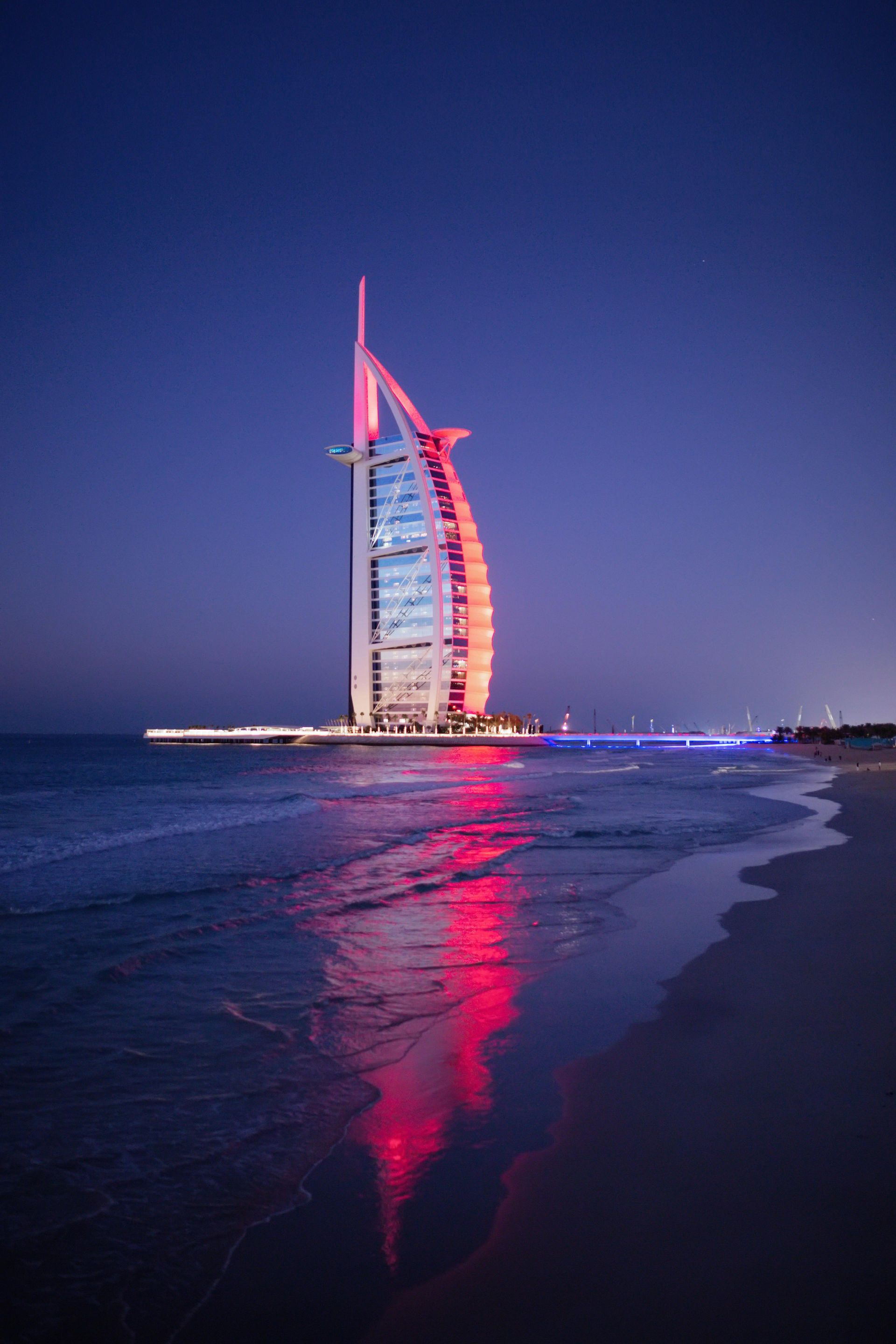 Burj Al Arab hotel lit red at dusk, reflecting on calm beach water