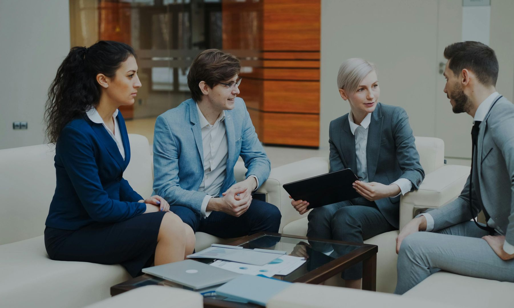 Four professionals in suits discuss documents in a bright office lounge.
