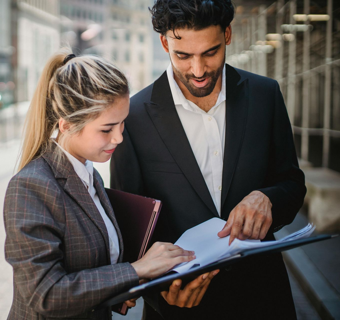 Two colleagues reviewing documents outdoors, one pointing at a page on a clipboard.