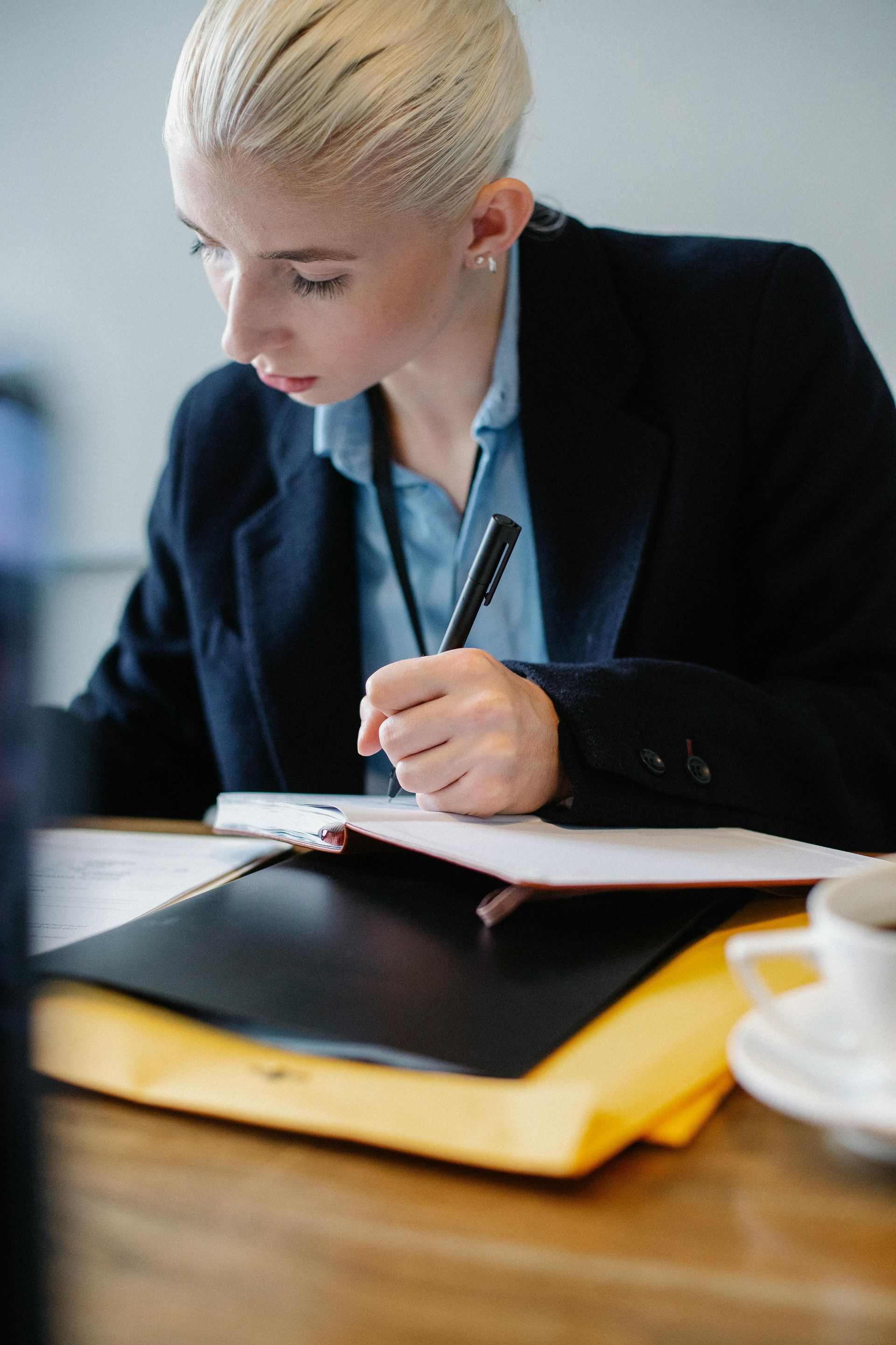 Person writing in a notebook at a desk with a black folder and coffee cup nearby