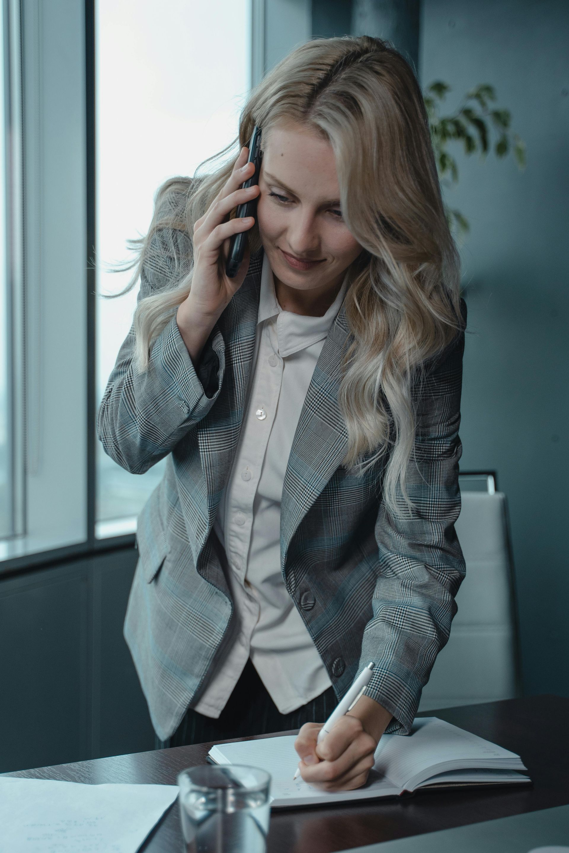 Woman on phone leaning over a desk, writing in a notebook in a modern office.