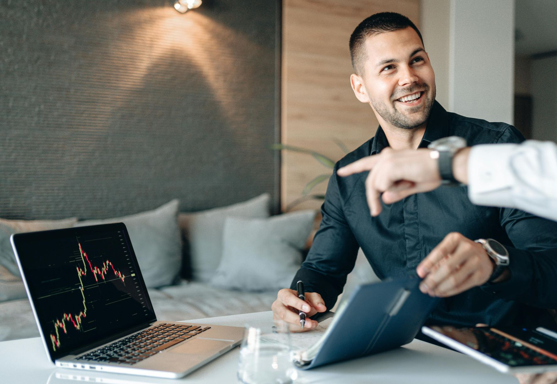 Man smiling at a table with a laptop, tablet, and coffee while another person points at the screen