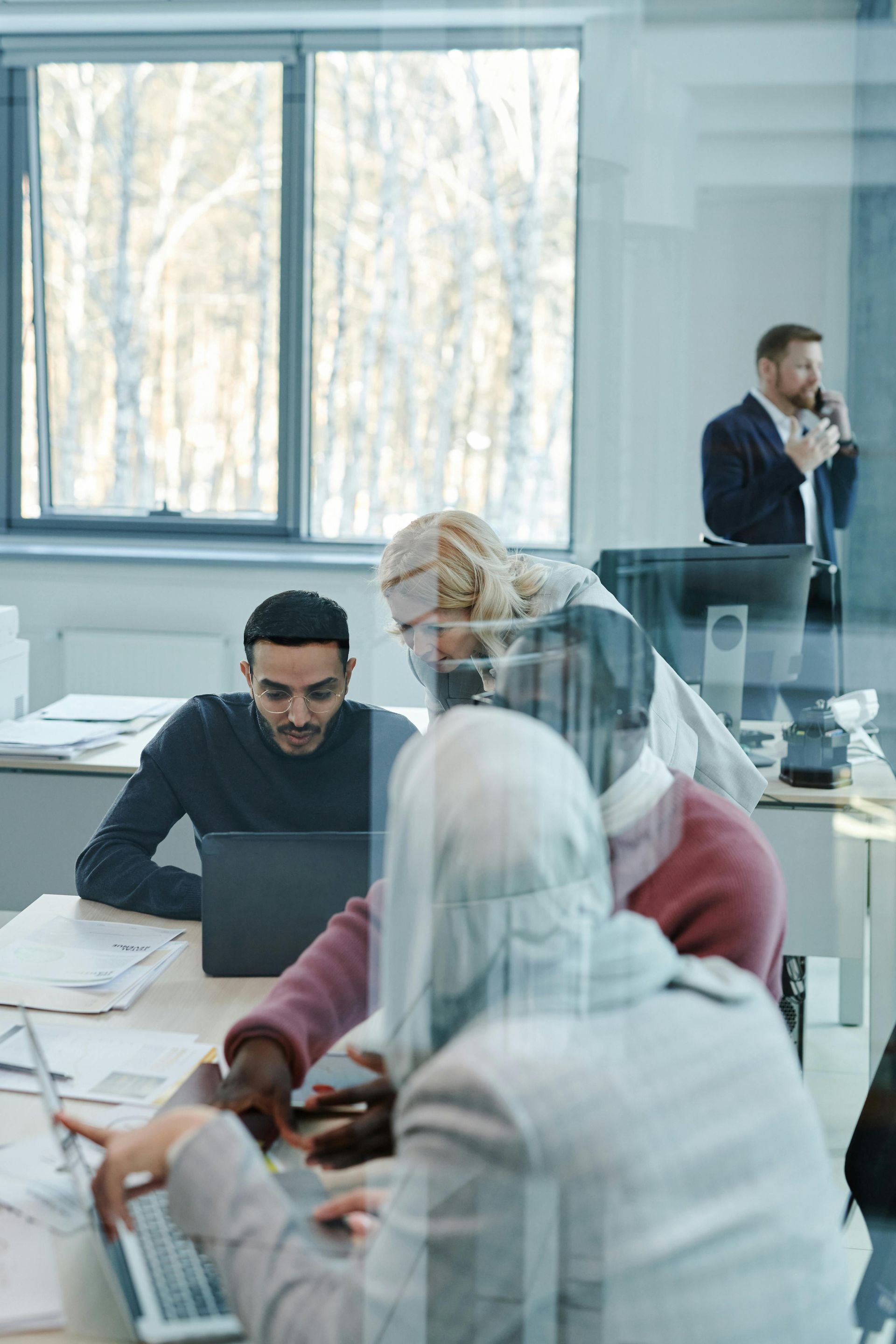 Office meeting with people working at desks; one person talks on a phone by the window.