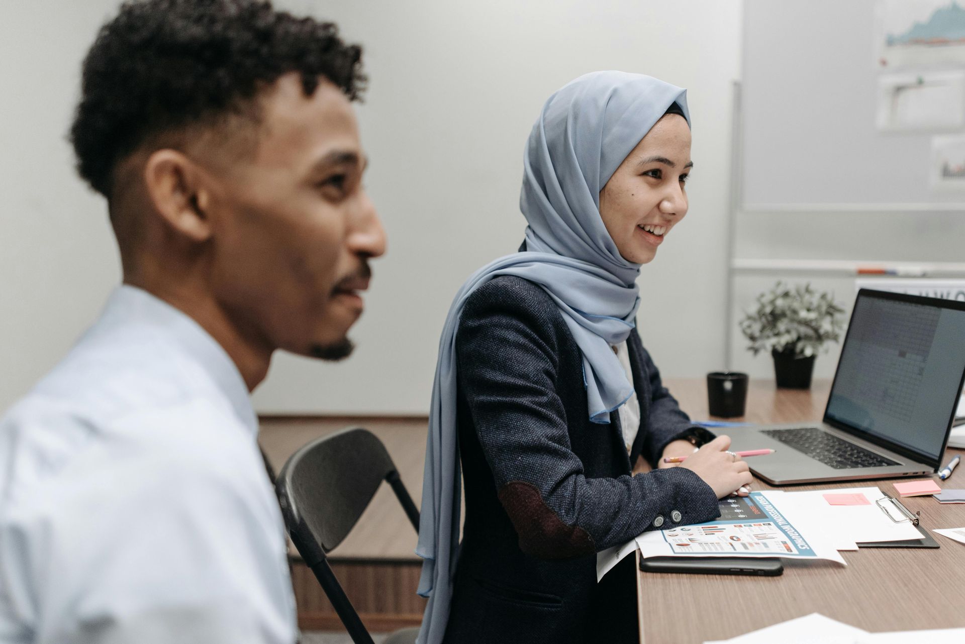 Two coworkers talking at a desk, with one using a laptop in a bright office