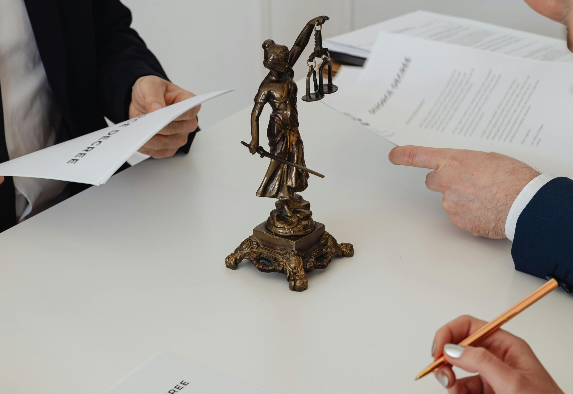 Hands exchanging documents around a bronze Lady Justice statue on a white table