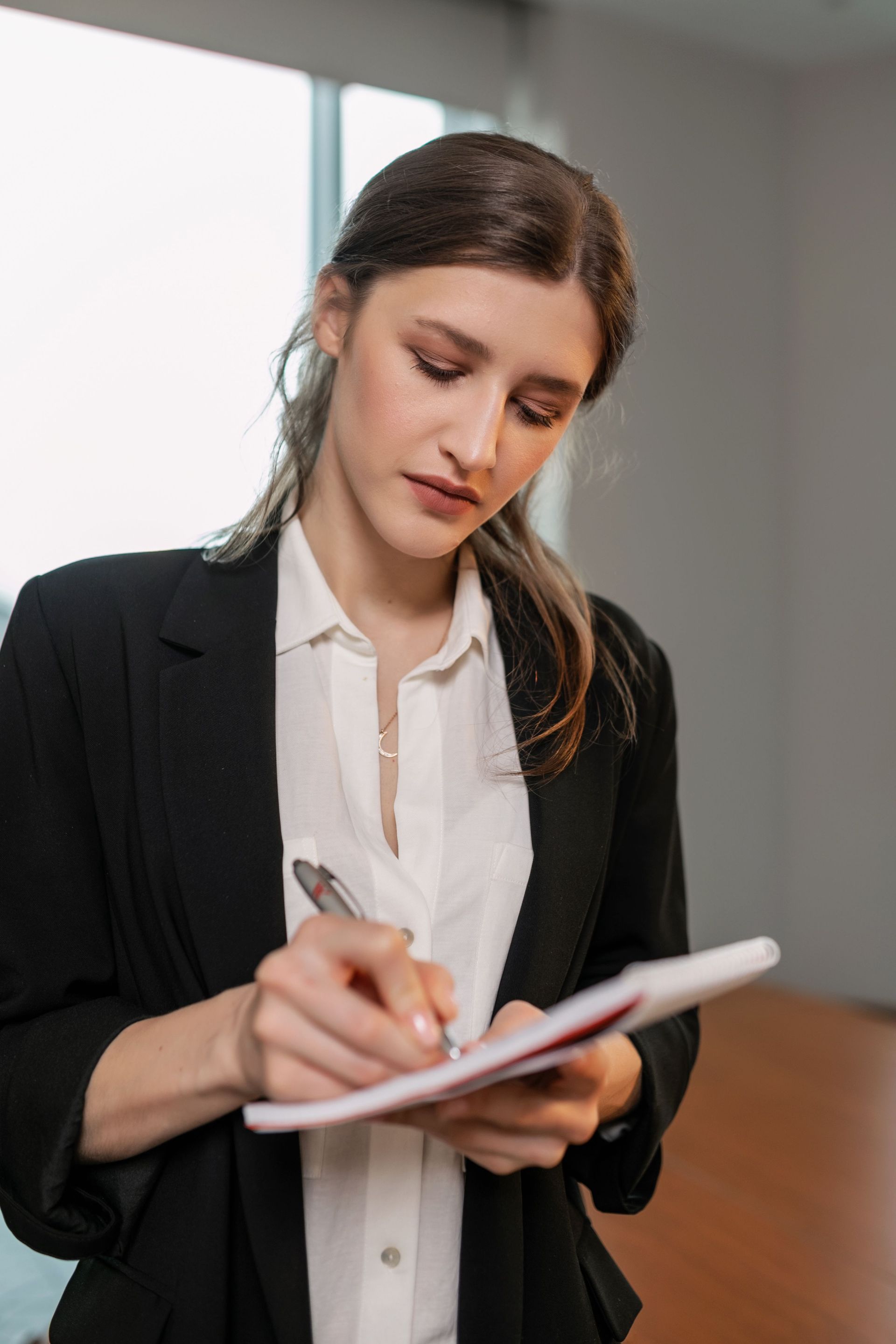 Woman on phone leaning over a desk, writing in a notebook in a modern office.