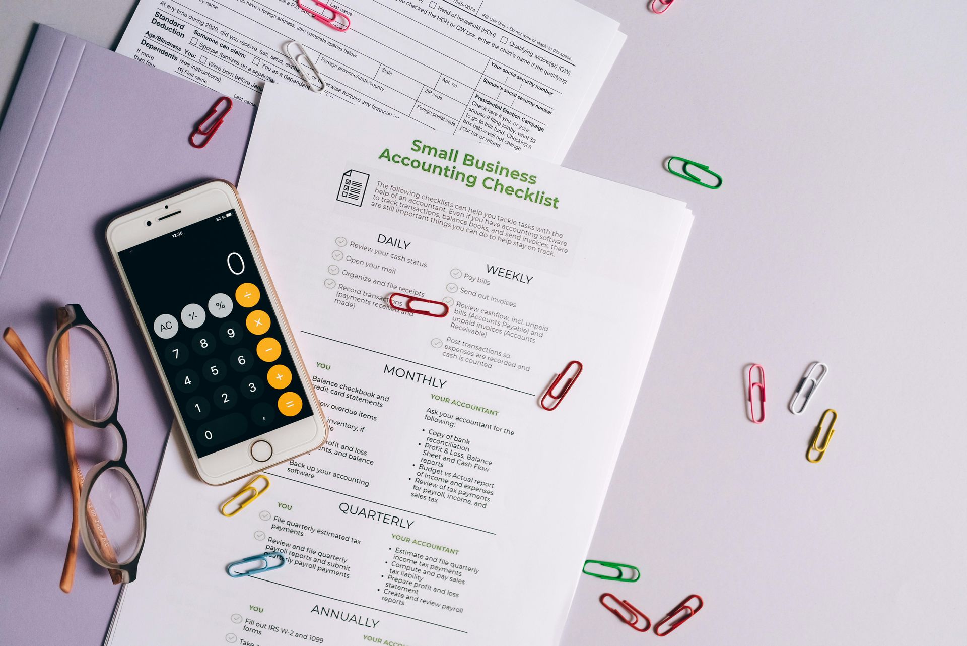 Desk with calculator, glasses, and scattered papers held together with colorful paper clips.