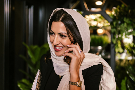 Woman in a white headscarf smiling while talking on a phone in a dimly lit café.